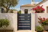 A dark gray aluminum garden gate is shown in front of a white house with a tiled roof, surrounded by greenery and flowering bushes. Produktbild-Vorschau