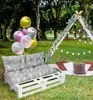 An outdoor picnic setup with a pallet sofa, cushions, balloons, and a teepee. Produktbild-Vorschau