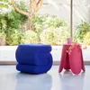 A blue pouf and a pink side table with a mug and flowers are placed in front of a large window with a garden view. Produktbild-Vorschau