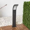 A dark gray LED bollard light with a motion sensor is placed on a wooden deck, surrounded by white pebbles and a green hedge. A white wall is in the background. Produktbild-Vorschau