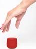 A hand with red nail polish is reaching down towards a small red Bluetooth speaker. Produktbild-Vorschau