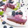 A slice of blueberry cheesecake with whipped cream and blueberries on a white plate, with a whole cheesecake in the background. Produktbild-Vorschau