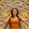 A woman wearing headphones lies on a sandy beach, enjoying an audiobook. Produktbild-Vorschau