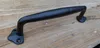 A black cast iron handle lies on a wooden surface. Produktbild-Vorschau