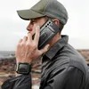 A man wearing a cap and jacket is on the phone outdoors, with a rugged landscape in the background. Náhled obrázku produktu