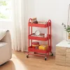 A red three-tiered rolling cart filled with various items, placed next to a sofa and a cabinet. Produktbild-Vorschau