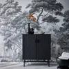 A black ribbed cabinet with two doors stands against a black and white forest-themed wallpaper. Orange flowers in a black vase are on top of the cabinet, along with a small bowl. A black side table with a book is to the right. Produktbild-Vorschau