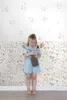 A young girl dressed in a denim romper stands in front of a floral wallpaper, holding a stuffed toy. Podgląd zdjęcia produktu
