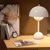 A white mushroom-shaped table lamp sits on a wooden desk next to a stack of books and an open magazine. Produktbild-Vorschau