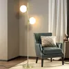 A modern floor lamp with two glass globes stands next to a dark green armchair with a textured cushion. Náhľad obrázku produktu