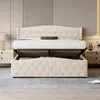 A beige upholstered storage bed with its hydraulic lift mechanism open, revealing storage space underneath. The bed is set in a bedroom with nightstands and lamps on either side. Náhľad obrázku produktu