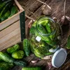 A rustic scene of cucumbers being prepared for pickling, with a wooden crate, a glass jar filled with cucumbers and dill, and garlic cloves scattered on a wooden surface. Podgląd zdjęcia produktu