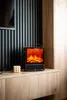 A small electric fireplace is placed on a wooden shelf, with a candle to its left and a black slatted wall behind it. Náhled obrázku produktu