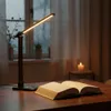 A desk lamp illuminates an open book on a wooden desk, with a candle and window in the background. Podgląd zdjęcia produktu