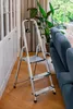 A silver step stool is placed on a wooden floor next to a blue sofa and a large potted plant, with curtains in the background. Anteprima dell’immagine prodotto