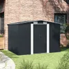 A dark gray metal garden shed is shown in a backyard setting, with a brick wall and a window visible behind it. The shed has white trim around the double doors and two ventilation grilles on the front gable. Produktbild-Vorschau