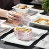 A person is placing a sandwich wrapped in paper onto a white plate. Other dishes are visible in the background. Produktbild-Vorschau