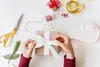 Hands are shown tying a white ribbon bow on a brown gift box, surrounded by gift wrapping supplies like scissors, ribbon, and sprigs of greenery. Podgląd zdjęcia produktu