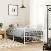 A bedroom scene with a white metal bed frame, bedding, a nightstand with a plant, a rug, and a clothing rack with clothes. Produktbild-Vorschau