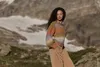 A woman stands in a mountainous landscape wearing a colorful sweater and tan pants. Produktbild-Vorschau