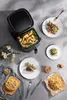 A black air fryer with food inside, surrounded by plates of food and cutlery on a gray surface. Produktbild-Vorschau