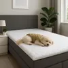 A small dog wearing a yellow vest sleeps on a white mattress topper on a bed. Produktbild-Vorschau