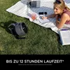 A woman is working on a laptop and writing in a notebook on a picnic blanket in the grass, with a portable fan nearby. Náhľad obrázku produktu