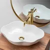 A white, petal-shaped vessel sink with a gold faucet is displayed on a wooden surface, with its reflection visible in a round, gold-rimmed mirror. Produktbild-Vorschau