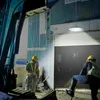 Two construction workers in a dimly lit industrial building, one operating machinery and the other standing near a large open garage door. Produktbild-Vorschau