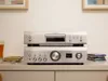 A silver Denon CD player and amplifier are stacked on a white surface, with CDs and a glass of drink to the side. Produktbild-Vorschau