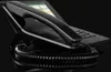 A close-up, angled shot of a black VoIP desk phone with a coiled cord resting on a reflective black surface against a black background. Produktbild-Vorschau