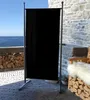 A black screen divider stands on a patio with a view of the ocean and a stone building in the background. Produktbild-Vorschau