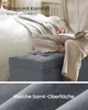 A person is sitting on a grey upholstered storage ottoman next to a bed, reading a book. The image is split into two sections, with the bottom section showing a close-up of the ottoman's fabric. Produktbild-Vorschau