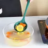 A person is using a green ladle to scoop soup with corn and carrots from a glass bowl. A black apron is visible. Náhled obrázku produktu