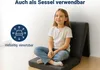 A young girl is sitting cross-legged on a folded mattress, which is set up as a chair. The background is a plain wall and floor. Produktbild-Vorschau