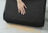 A hand is shown touching a dark gray doormat that is partially rolled up, revealing some dirt or sand underneath it on a light gray tiled floor. Podgląd zdjęcia produktu