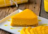 A slice of mango cheesecake on a white plate, garnished with mango puree and sliced mangoes, on a wooden table. Podgląd zdjęcia produktu