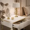 A close-up shot of a white high-gloss nightstand with gold accents, featuring a lamp, a bust, and an open drawer with a book inside. Náhled obrázku produktu