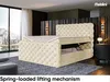 A luxurious beige velvet bed with a tufted headboard and a lifted mattress revealing storage space, set in a modern bedroom with a large window overlooking a mountain landscape. Produktbild-Vorschau
