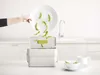 A hand is shown pouring green beans from a plate into a compost bin on a kitchen counter. Náhled obrázku produktu