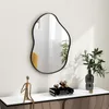 A modern, irregularly shaped mirror hangs on a white wall above a console table. The table holds decorative items including a metallic figurine, a stack of books with a geometric sculpture on top, and a vase with greenery. The mirror reflects a yellow chai Produktbild-Vorschau