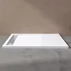 A white rectangular shower tray with a silver drain is shown on a concrete floor against a brown wall. Produktbild-Vorschau