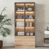 A bathroom cabinet with glass doors, illuminated shelves displaying towels and toiletries, and two drawers at the bottom. A plant is visible to the left. Voir les images du produit