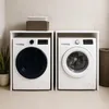 Two washing machines are shown side-by-side in a laundry room setting, with a shelf above holding folded towels and a basket. Voir les images du produit
