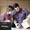 A father and son are working together in a workshop, measuring a wooden object with a tape measure. Náhled obrázku produktu