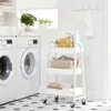 A white rolling cart with three shelves is shown in a laundry room setting next to two washing machines. Náhled obrázku produktu