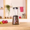 A blender is shown on a wooden countertop with some fruit in the foreground and shelves with jars and plants in the background. Produktbild-Vorschau