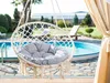 A hanging chair with a cushion is placed outdoors near a swimming pool, with lounge chairs and greenery in the background. Produktbild-Vorschau