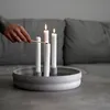 A hand is lighting a candle on a round, grey candle holder with four white candles, placed on a grey table in front of a grey sofa. Produktbild-Vorschau