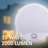 A close-up of a white LED outdoor wall light with a motion sensor, set against a blurred background of a house at dusk with illuminated windows. Produktbild-Vorschau